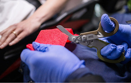Translation missing: fr.A person wearing blue gloves using the shears on a Woodland colored Leatherman Raptor Rescue to cut a piece of red medical fabric