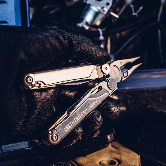 Translation missing: nl. A person wearing black gloves holding a Leatherman Wave+, showcasing it's pliers and tools inside the handle, getting ready to work on a vehicle