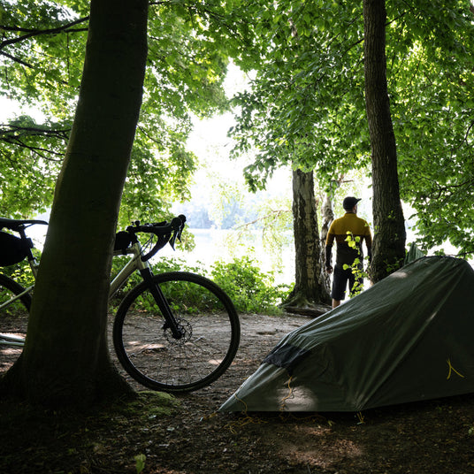 Translation missing: fr.Cyclist standing beside a tent and bicycle in a forest clearing near a lake