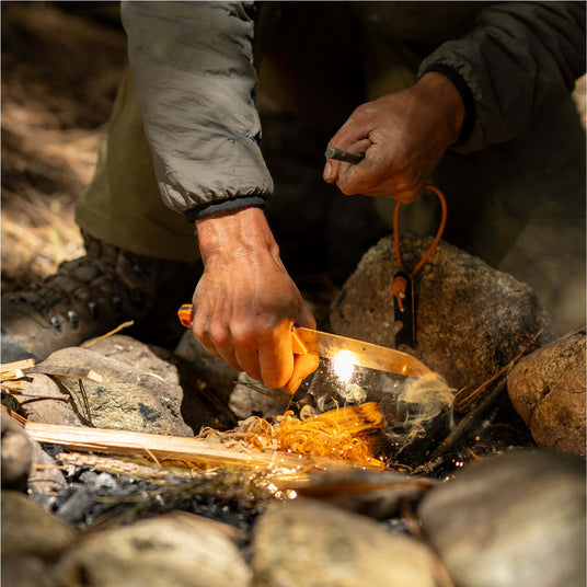 Translation missing: nl.Person using a Leatherman Pioneer knife and fire starter to ignite feather sticks in a campfire setup, shown in an outdoor setting with sparks and smoke rising from the tinder