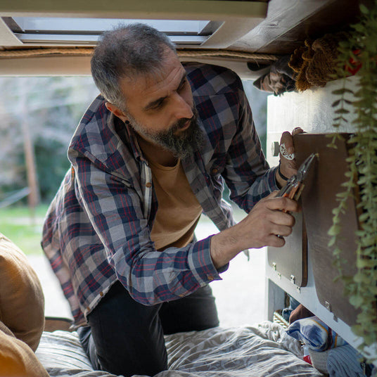 Translation missing: de.Person using a Leatherman Signal multi-tool to make repairs inside a camper van, kneeling on a bed while gripping the tool to tighten a fastener on a cabinet panel