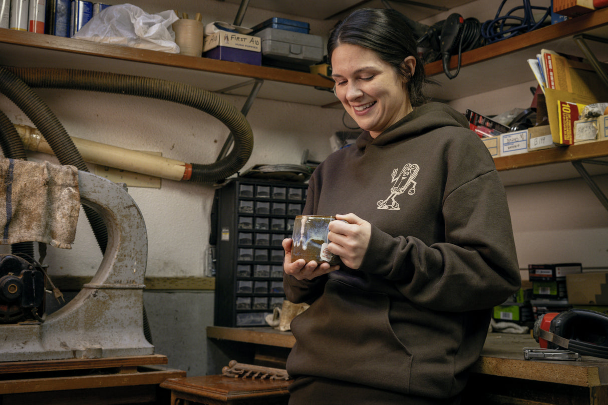 Translation missing: en.A person wearing a brown Leatherman Mr. Wave hoodie standing in a workshop holding a ceramic mug, smiling while surrounded by tools and equipment