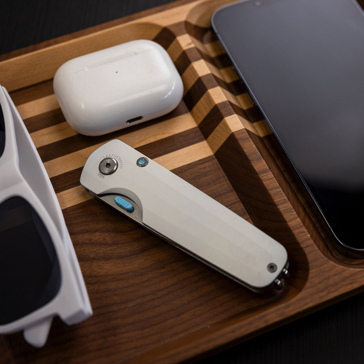 Translation missing: fr.Leatherman Blazer Alpine folding knife in closed position placed in a wooden valet tray alongside wireless earbuds, a smartphone, and sunglasses, highlighting everyday carry essentials