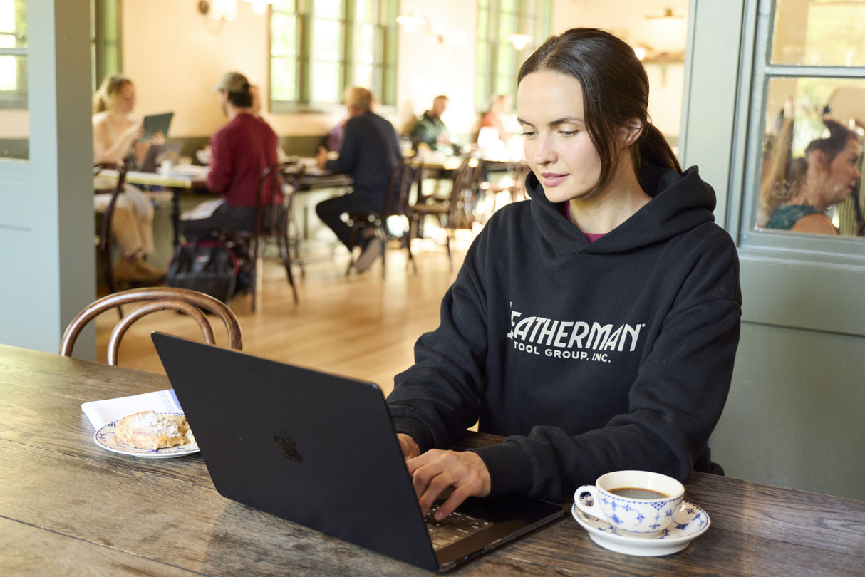 Translation missing: en.A woman sitting indoors working on a computer while wearing a black Leatherman Blade & Grain hoodie with a pastry and coffee on either side of the laptop
