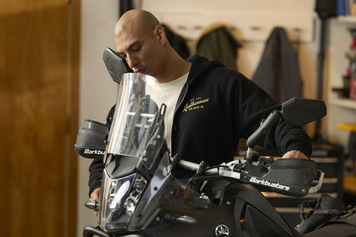 Translation missing: en.A person wearing a black Leatherman Vintage Zip-Up hoodie standing next to a motorcycle in a garage, with the hoodie’s gold Leatherman logo visible on the chest