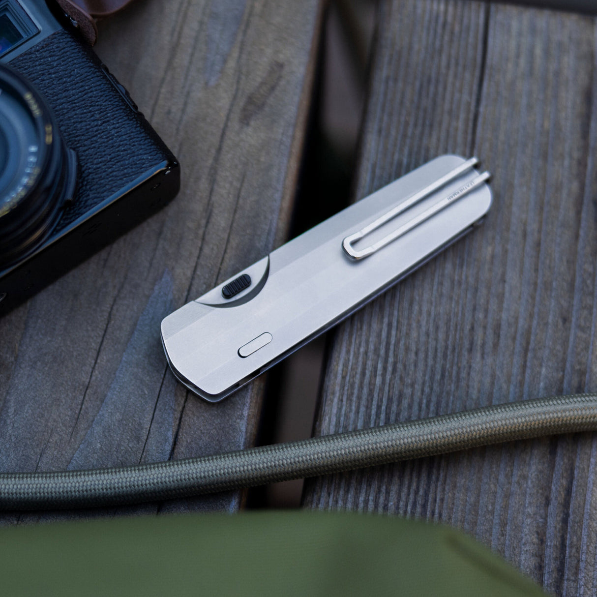 Translation missing: de.Leatherman Blazer Stainless Steel folding knife in closed position resting on a wooden surface beside a camera and outdoor gear, emphasizing everyday carry in an outdoor setting