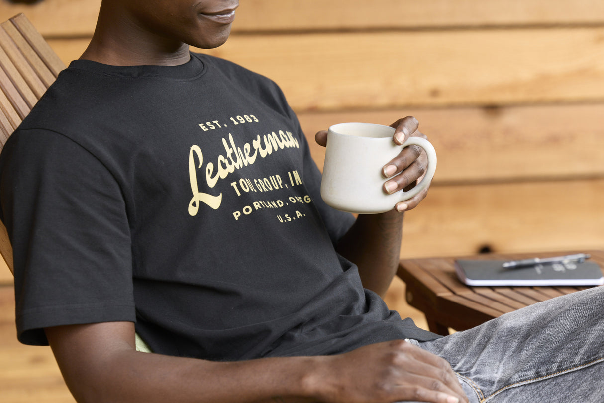 Translation missing: de.A person wearing a black Leatherman Vintage Tee relaxing on a wooden deck chair, holding a white mug with a notebook and pen on a small table nearby