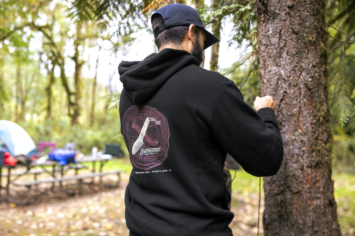 Translation missing: en.A person wearing a black Leatherman Blade & Grain hoodie standing next to a tree in a forest campsite. The hoodie’s back graphic shows a pocketknife illustration with “Leatherman Tool Group” text