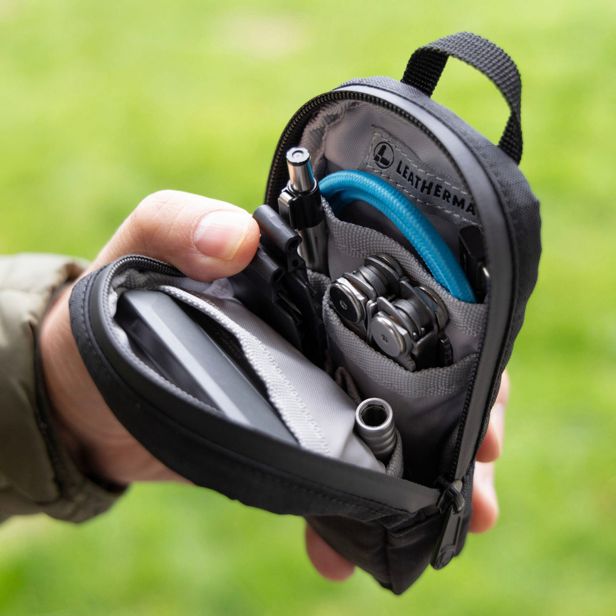 Translation missing: de.A hand holds an open black Leatherman Tool Pouch, revealing a neatly organized interior with a multi-tool, flashlight, pen, charging cable, and other everyday carry essentials. The pouch features a durable design with multiple compartments and a branded interior label