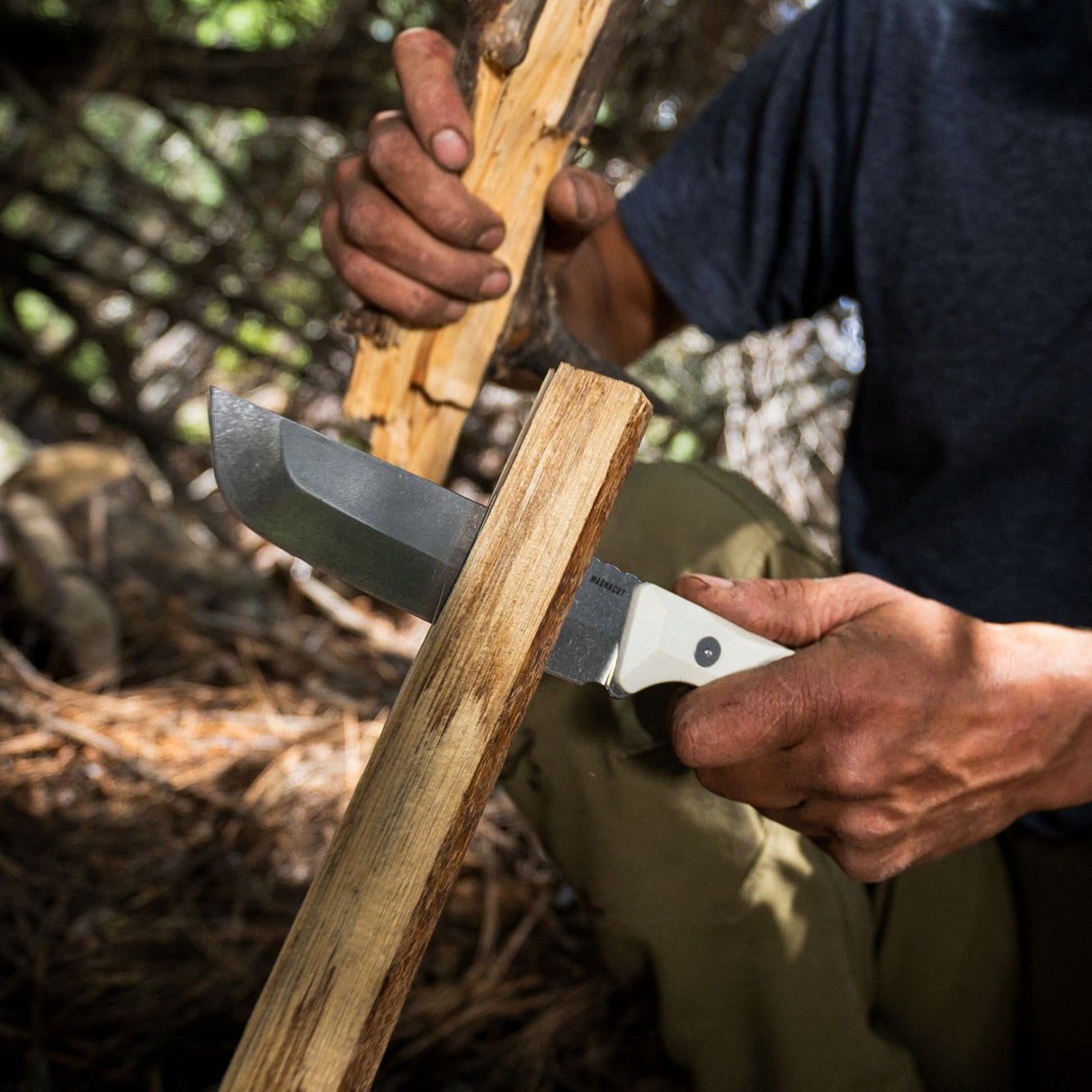 Translation missing: fr.A man skillfully uses a Leatherman Alpine Pioneer knife to slice through a wooden plank, demonstrating craftsmanship in a workshop environment