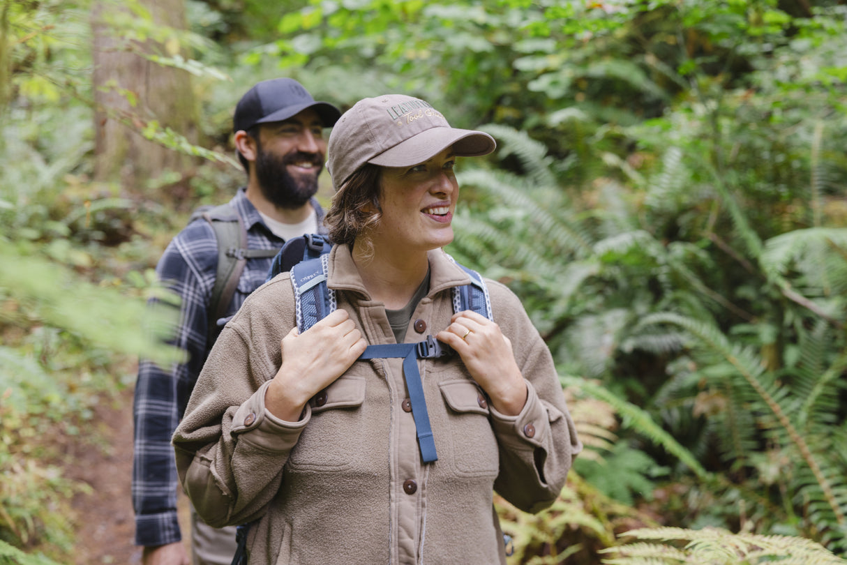 Translation missing: es.A woman wearing a Leatherman Dad Hat in Driftwood color hiking through a forest with a backpack smiling as another hiker follows behind