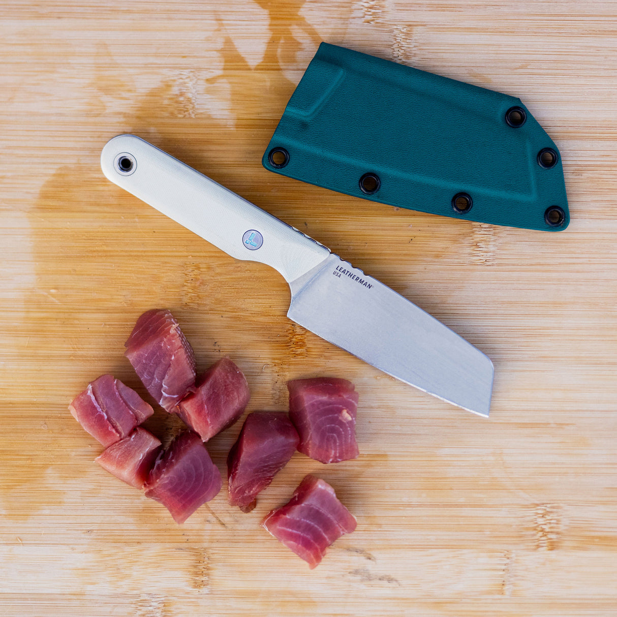 Translation missing: en.A Leatherman Rustle Alpine knife on a wooden cutting board next to a teal sheath and freshly cut chunks of raw fish