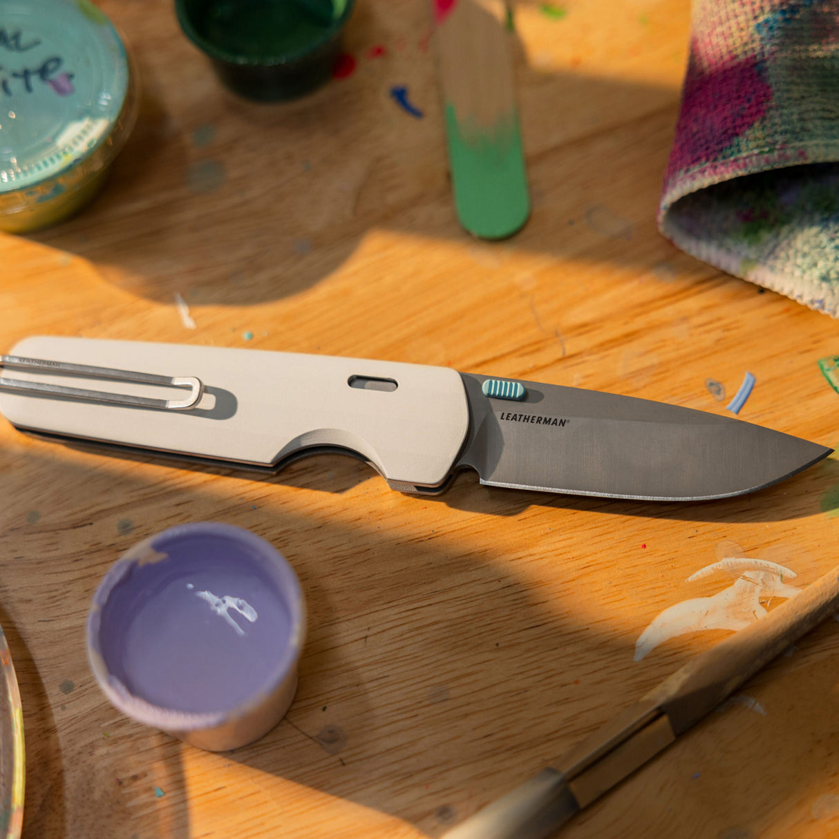 Translation missing: fr.Leatherman Blazer Alpine folding knife shown open on a wooden table surrounded by paint supplies and a brush, highlighting its role as a creative or everyday utility tool