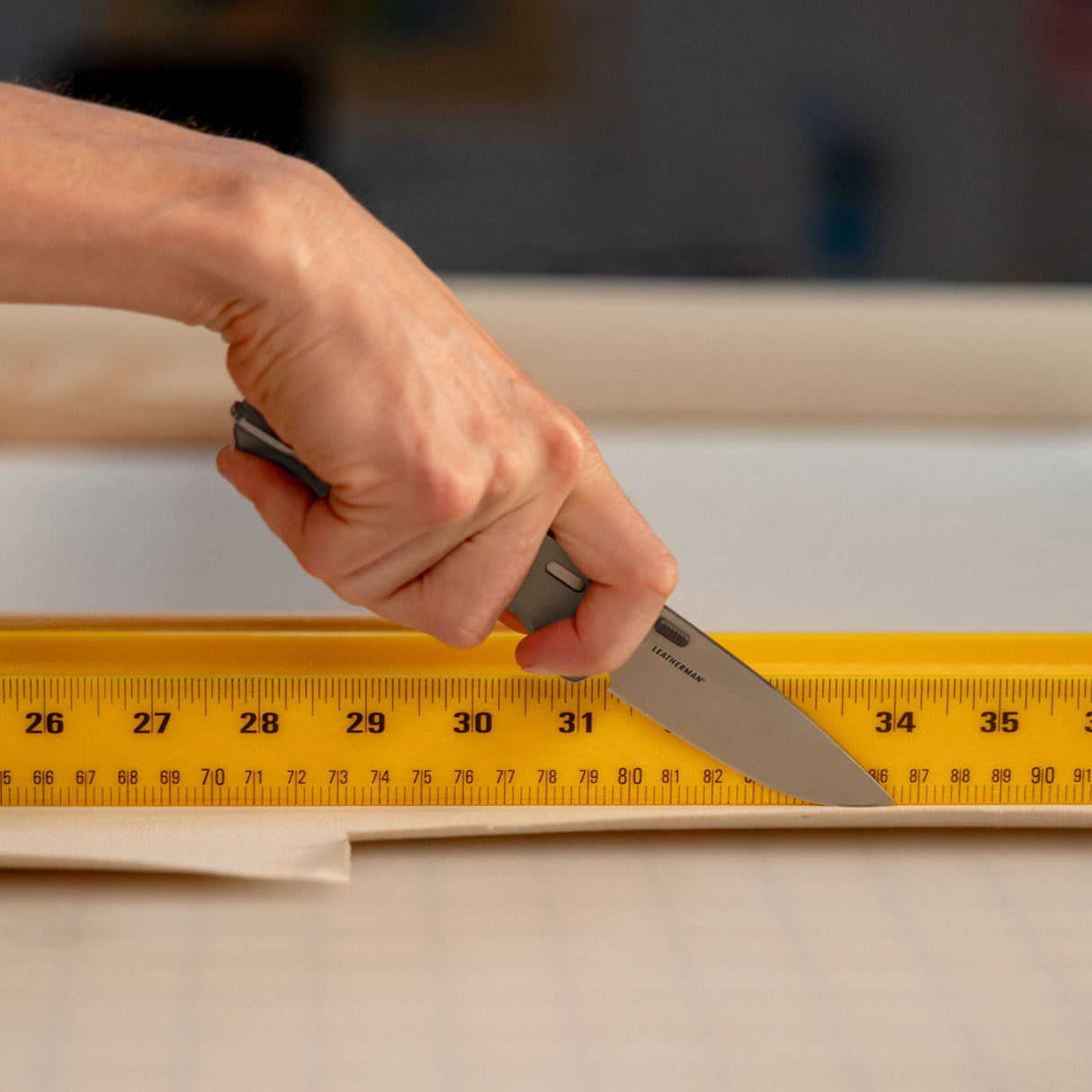 Translation missing: de.Person using a Leatherman Blazer Stainless Steel folding knife to cut fabric along a yellow measuring ruler on a gridded work surface, shown in a close-up of the hand and cutting action