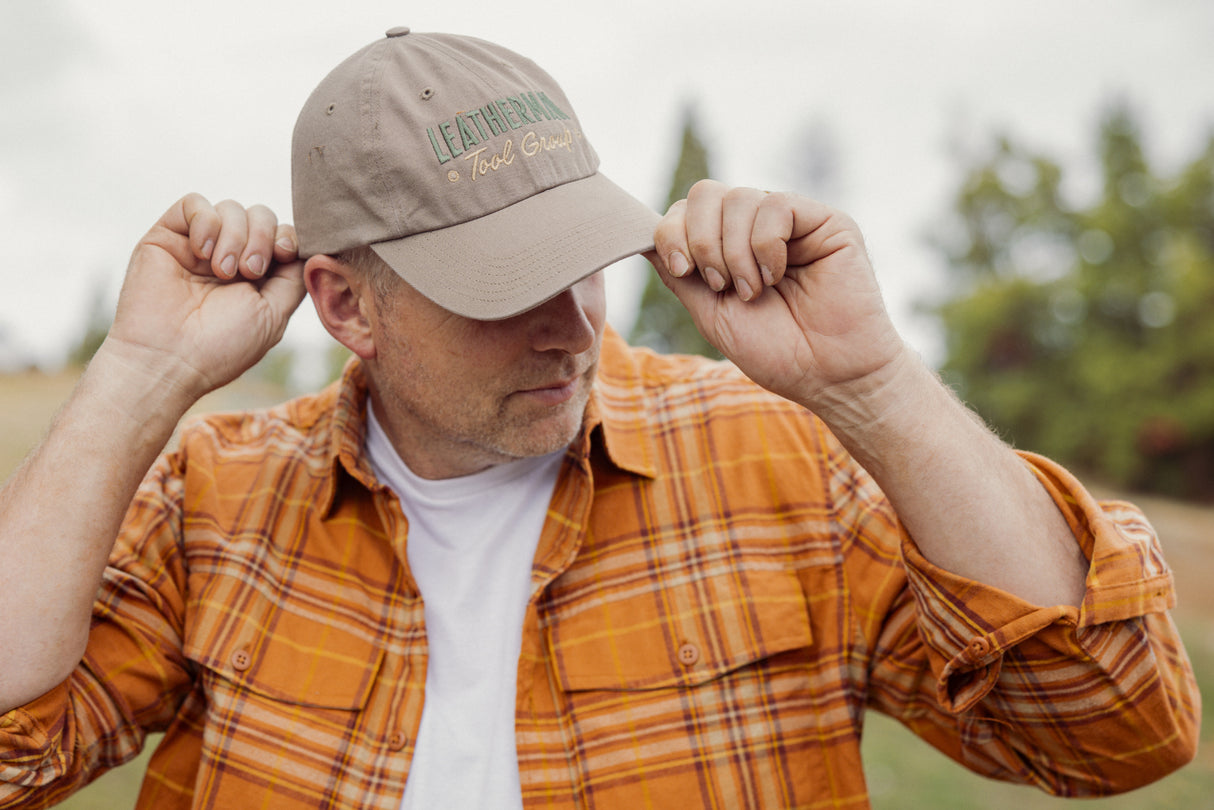 Translation missing: es.A person wearing a Leatherman Dad Hat in Driftwood color adjusting the brim while outdoors, dressed in an orange plaid shirt