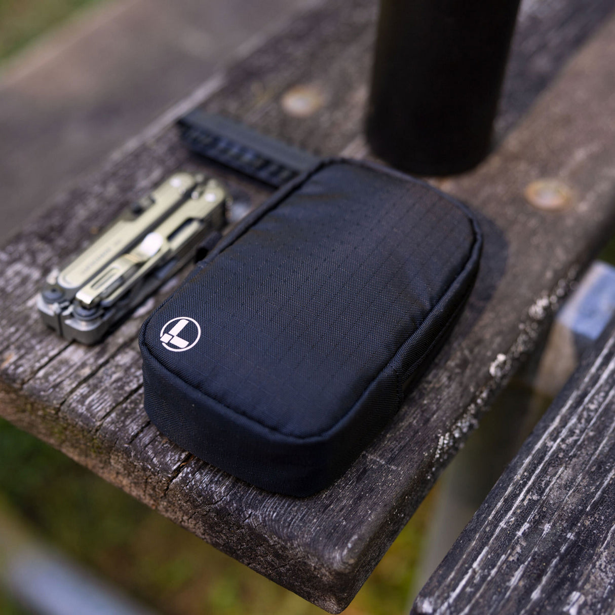 Translation missing: de.A black Leatherman Tool Pouch with a white logo rests on a weathered wooden bench outdoors, positioned beside a Leatherman ARC multi-tool and black water bottle, suggesting a rugged, ready-for-anything everyday carry setup