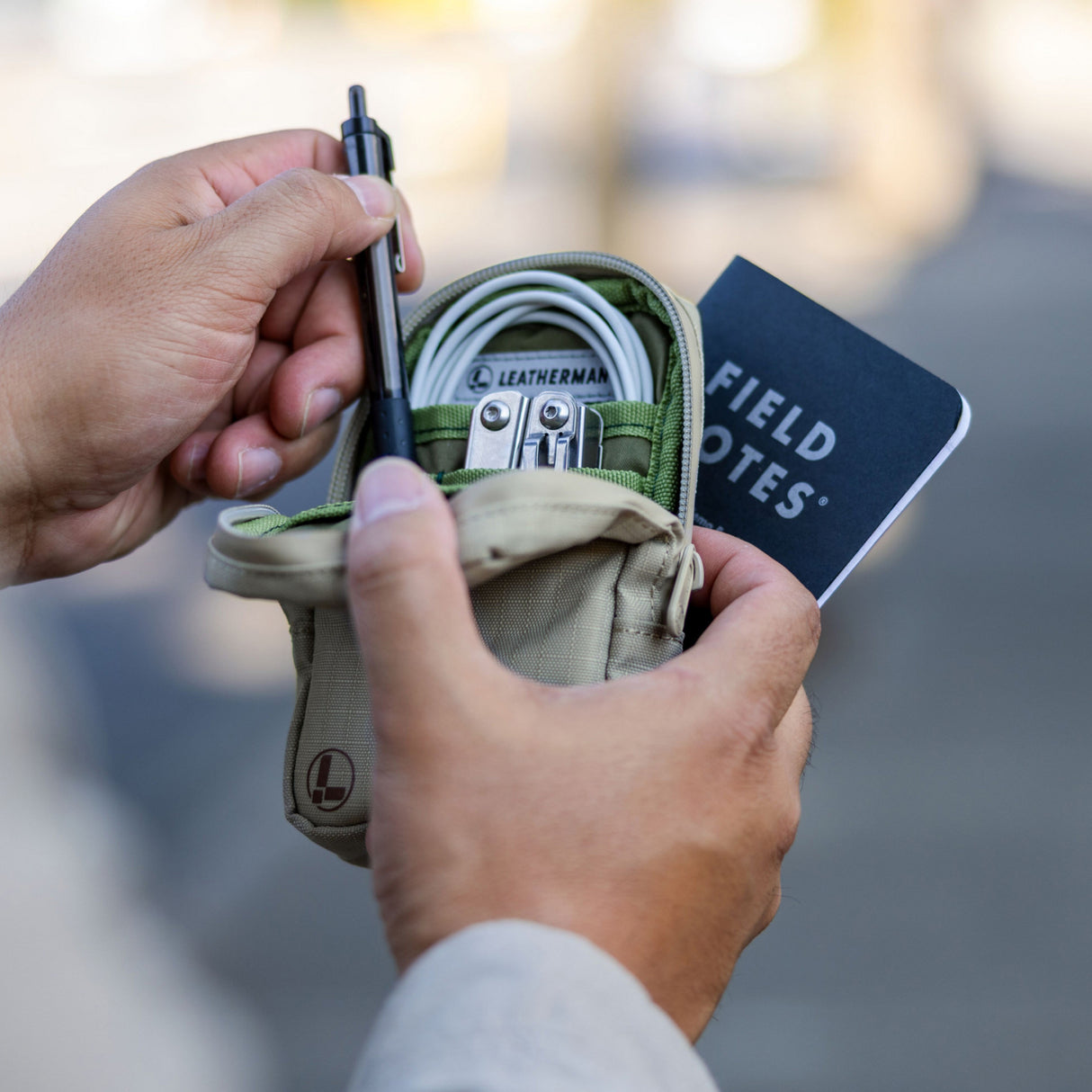Translation missing: nl.A person holds open a Leatherman Tool Pouch in Mossy Fog color, revealing neatly organized items including a Leatherman multi-tool, charging cable, pen, and Field Notes notebook—illustrating the pouch's utility for everyday carry and organization