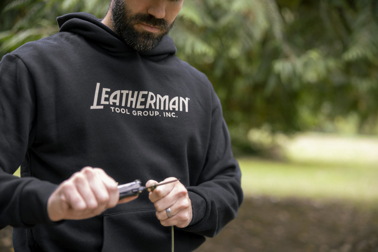 Translation missing: en.A person wearing a black Leatherman Blade & Grain hoodie using a Leatherman multitool to cut a piece of rope outdoors, showing the front logo of the hoodie that reads “Leatherman Tool Group, Inc.”