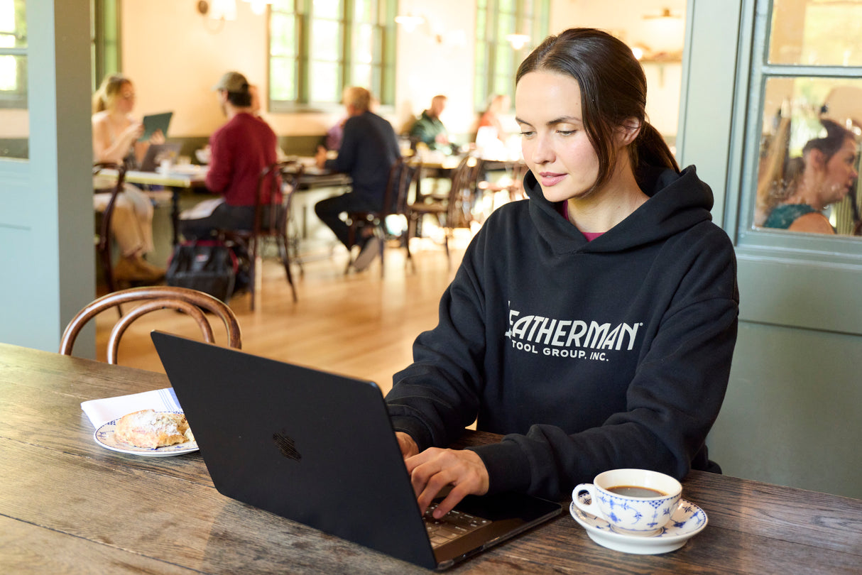 Translation missing: en.A woman sitting indoors working on a computer while wearing a black Leatherman Blade & Grain hoodie with a pastry and coffee on either side of the laptop
