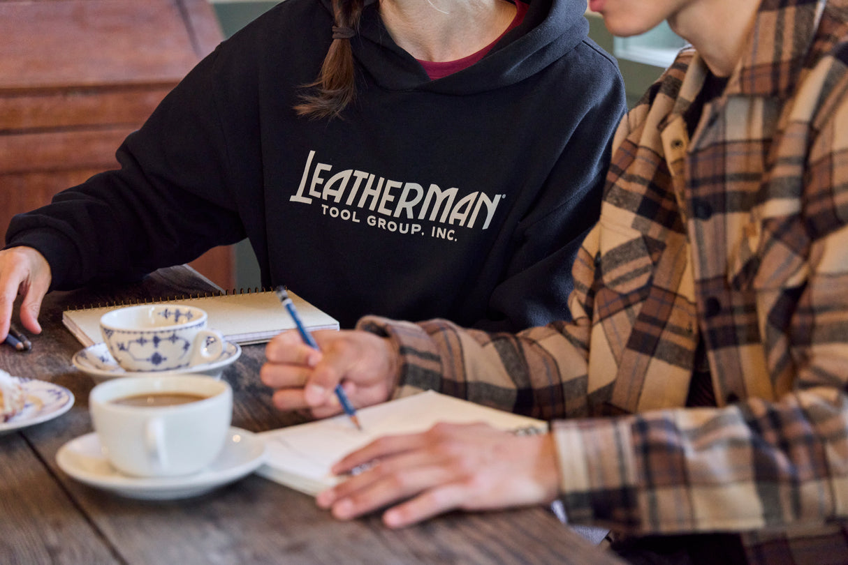 Translation missing: en.Two people sitting at a table with coffee, pastries and notebooks. One is wearing a black Leatherman Blade & Grain hoodie with 'Leatherman Tool Group Inc.' on the front