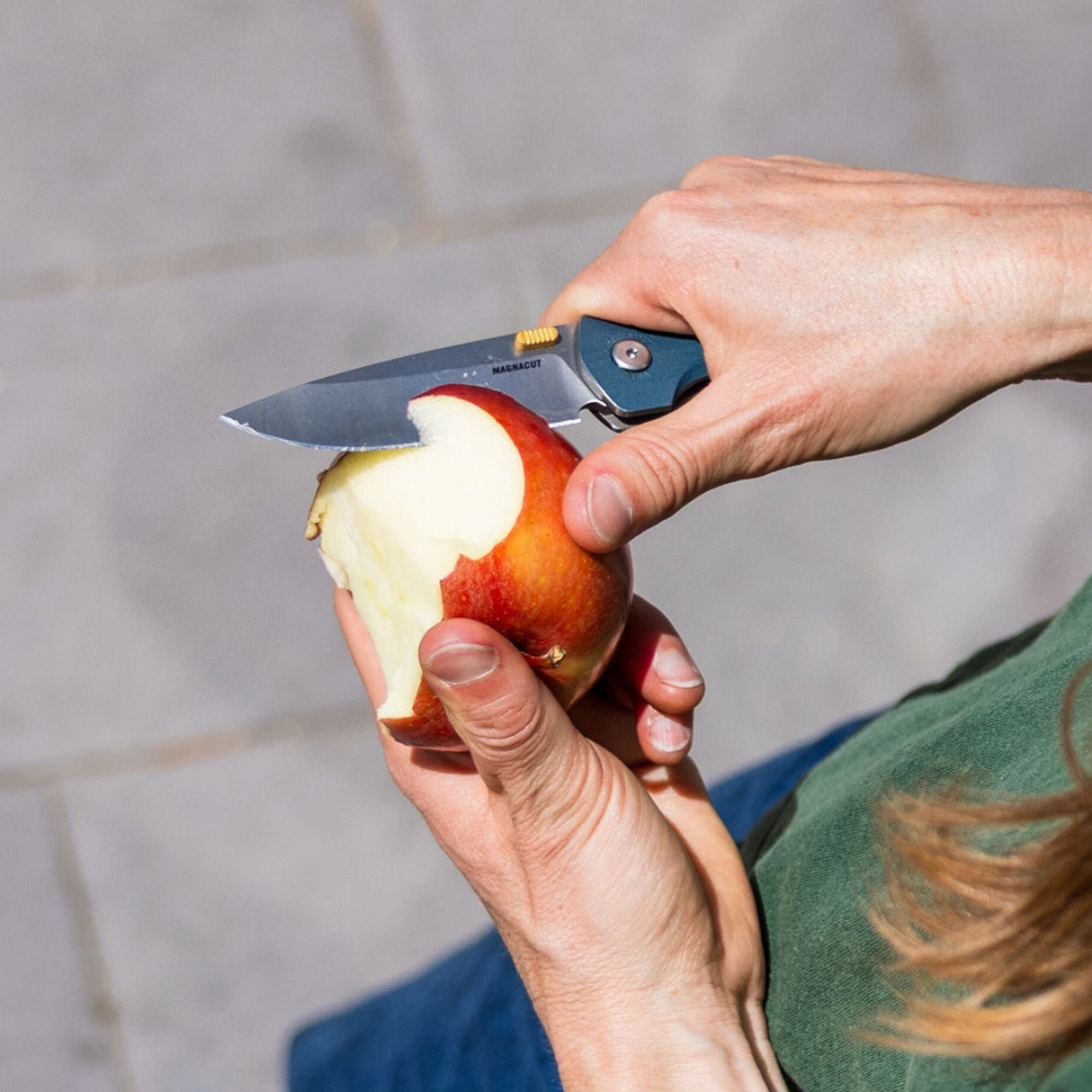 Translation missing: nl.Person using a Leatherman Blazer Denim folding knife to peel an apple outdoors, shown in a close-up view of the hands and knife in use