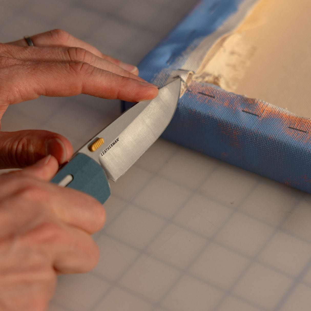 Translation missing: nl.Person using a Leatherman Blazer Denim folding knife to cut canvas material from a wooden frame, shown in a close-up on a gridded surface