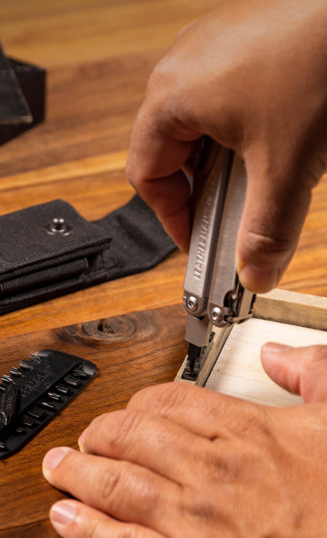 Translation missing: de.Someone using a Leatherman Wave bit to screw the backing on a wooden frame with a bit kit and a Leatherman black nylon sheath with pockets laying next to the wooden frame on a wooden table