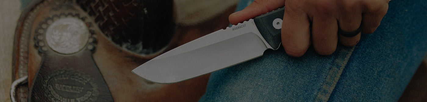 Translation missing: nl.Close-up of a person holding a Leatherman fixed-blade knife with "MAGNACUT" engraved on the blade, positioned over their denim-clad leg next to a leather saddle