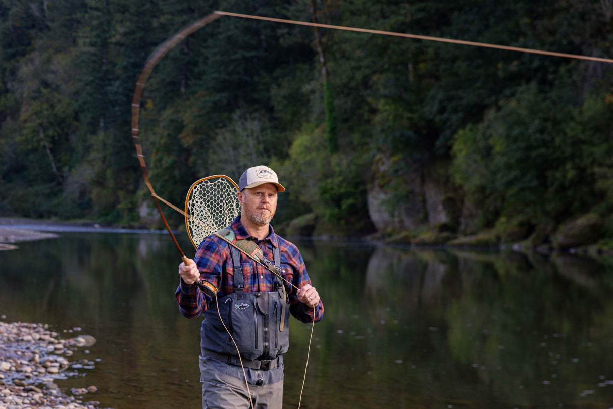 Translation missing: de.A person wearing a beige Leatherman PNW Trucker hat fly-fishing in a river surrounded by trees