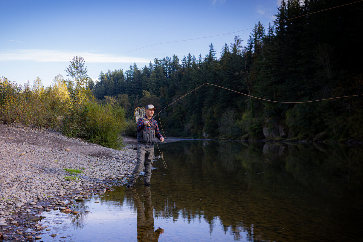 Translation missing: de.A person wearing a beige Leatherman PNW Trucker hat fly-fishing in a river surrounded by trees
