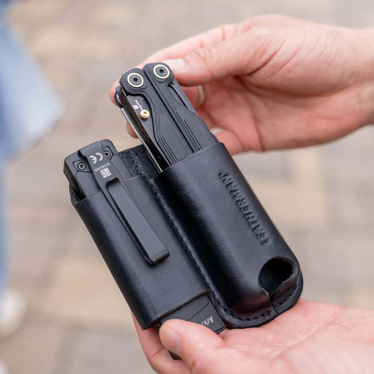 Translation missing: fr.Hands holding a black leather sheath carrying two black multitools outdoors