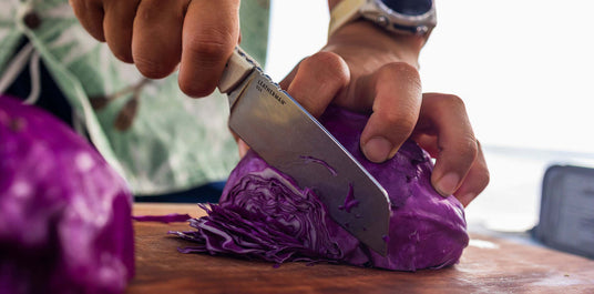 Translation missing: de.Person using a Leatherman Rustle knife to slice a head of purple cabbage on a wooden cutting board, shown in a close-up with focus on the blade and hands in action