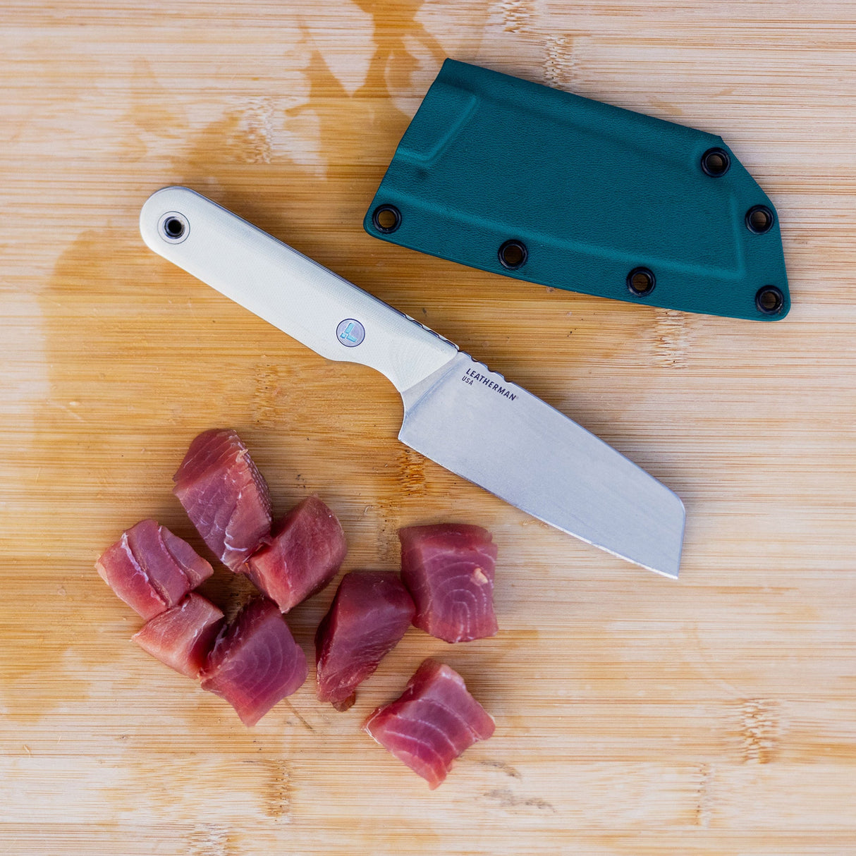 Translation missing: de.A Leatherman Rustle Alpine knife on a wooden cutting board next to a teal sheath and freshly cut chunks of raw fish