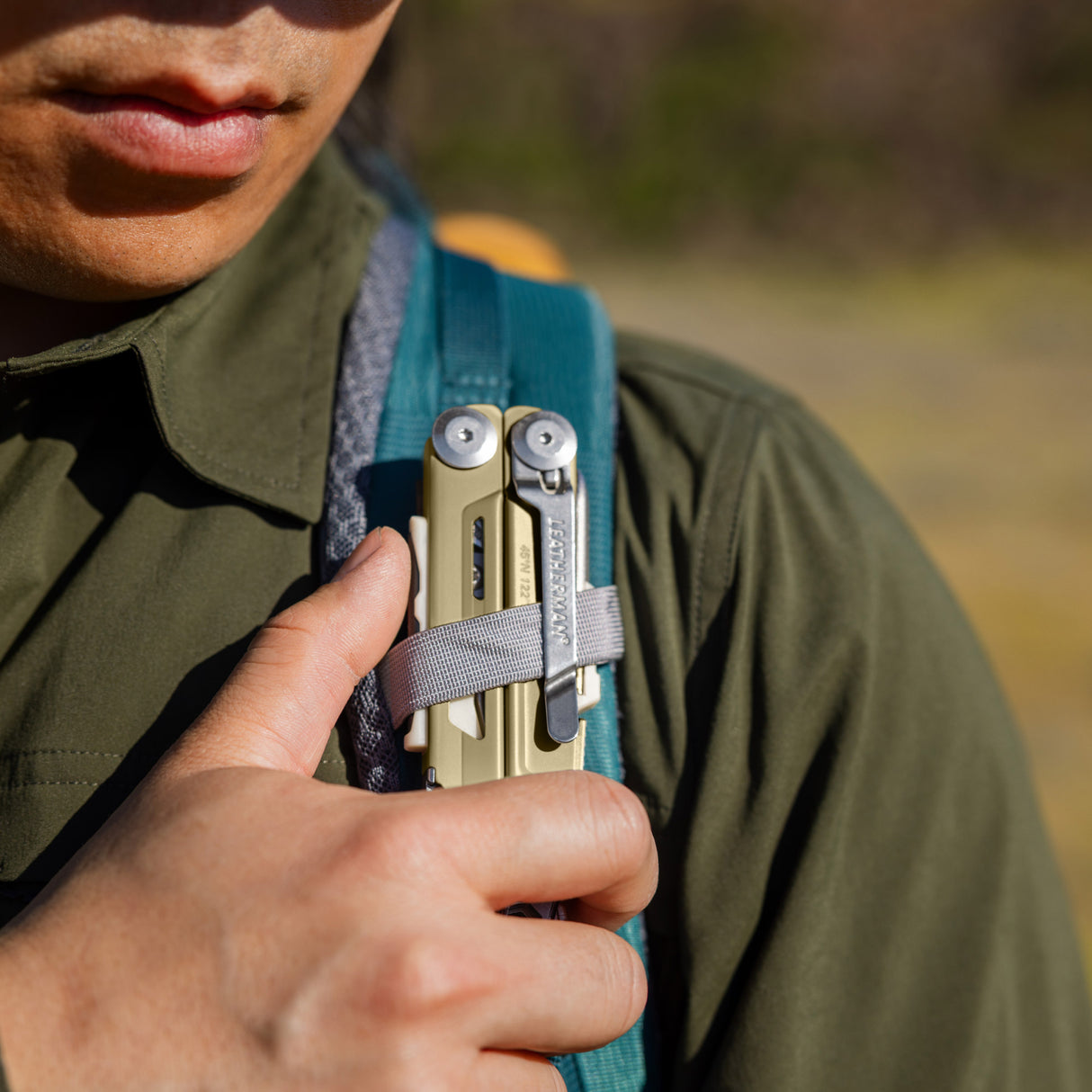 Translation missing: es.Person securing a Sandstorm multitool to a backpack shoulder strap with a gray loop outdoors