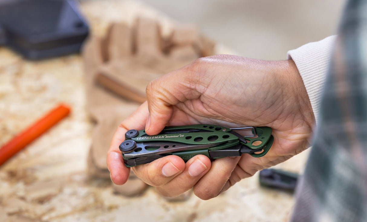 Translation missing: es.Person holding a closed OD Green Leatherman Skeletool® CX multitool over a wooden work bench