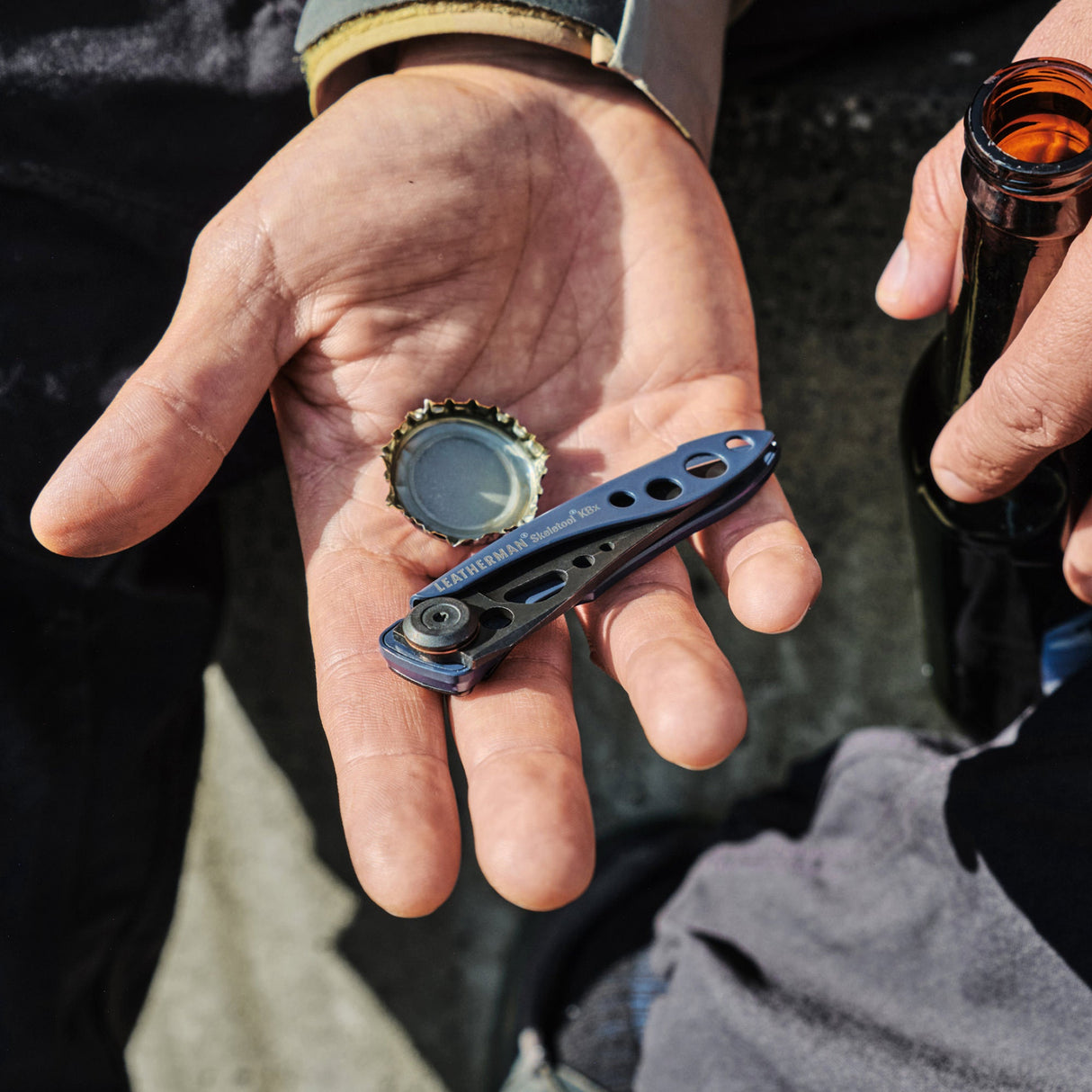 Translation missing: pl.Hand holding a blue Skeletool KBX and bottle cap next to a person gripping an open glass bottle