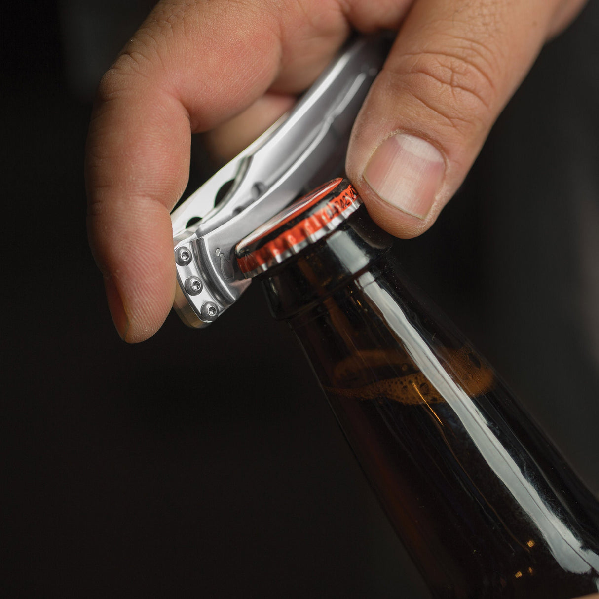 Translation missing: pl.Hand using a stainless steel Skeletool KBX to open a capped glass bottle