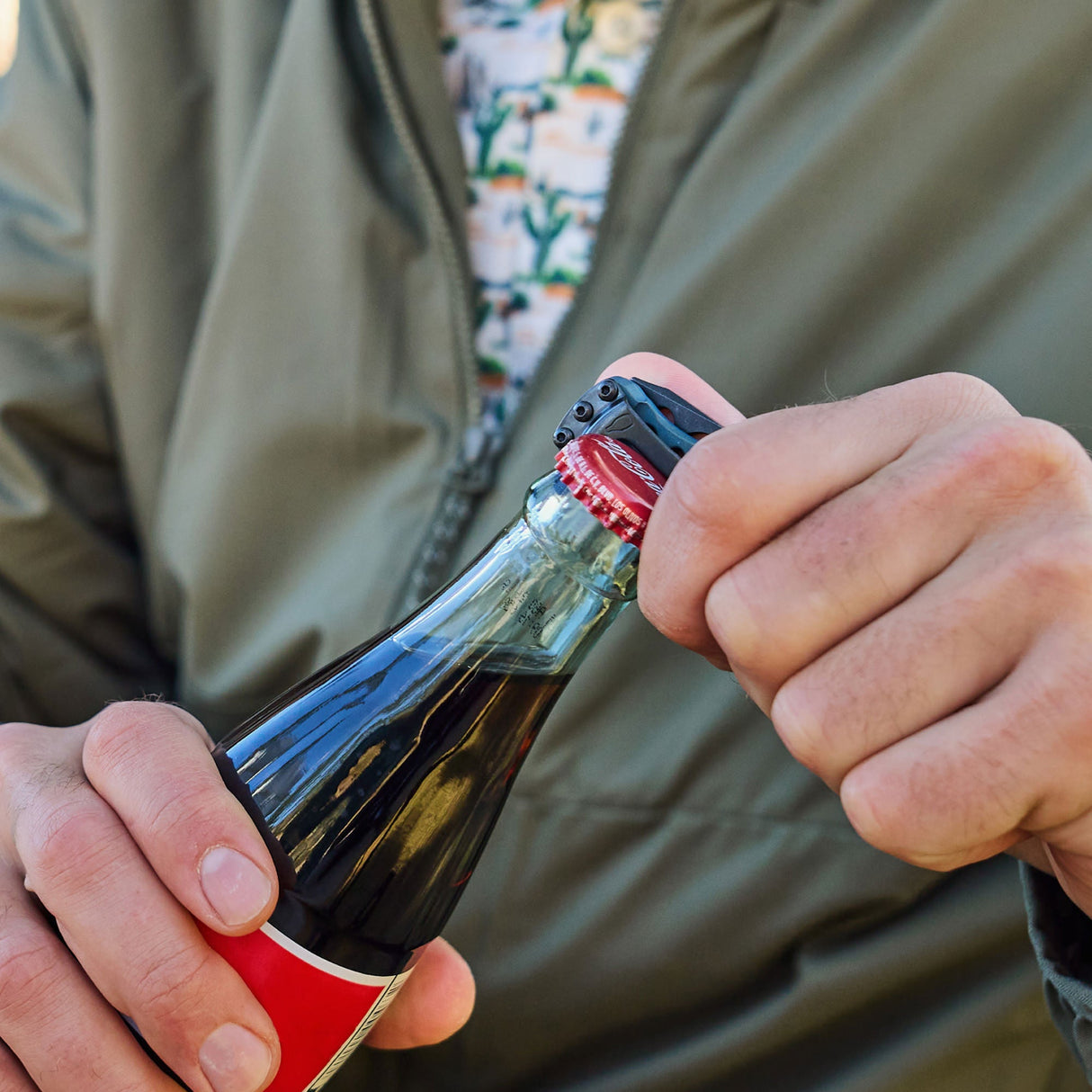 Translation missing: pl.Hand using a Skeletool KBX to pry off the cap of a glass soda bottle