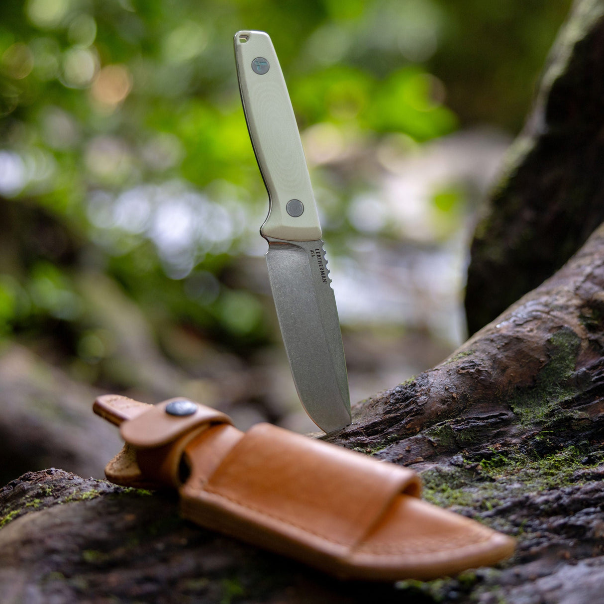 Translation missing: de.Leatherman Trac Alpine fixed-blade knife embedded in a mossy tree trunk in an outdoor setting, with a tan leather sheath resting in the foreground