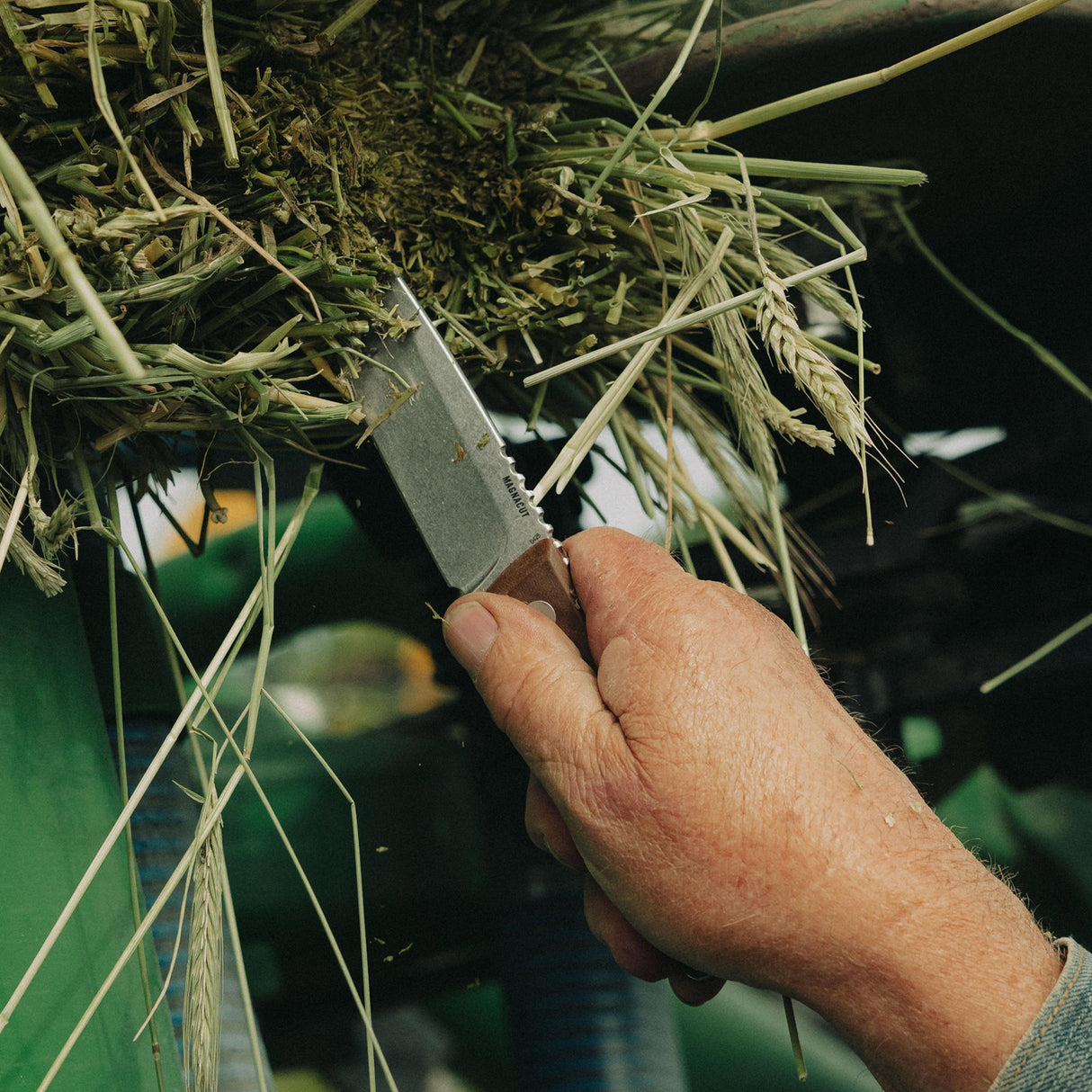 Translation missing: en.Person using a Leatherman Trac Earth Brown fixed-blade knife to clear or cut through compacted straw and plant debris from agricultural equipment in a farm setting