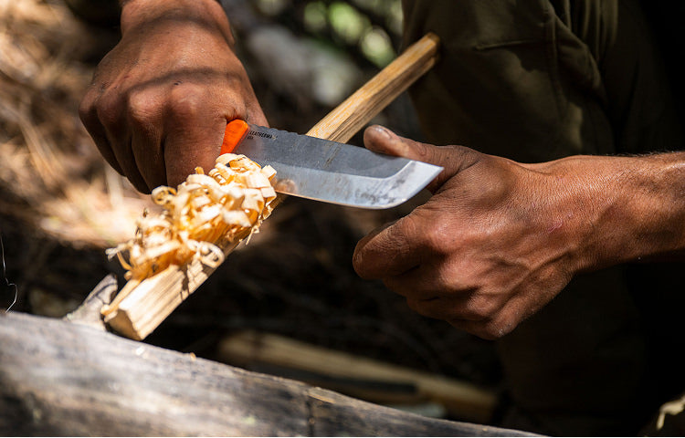 Translation missing: nl.Close-up of a person using a Leatherman knife with an orange handle to carve wood shavings from a stick, surrounded by natural outdoor elements, suggesting a bushcraft or survival setting