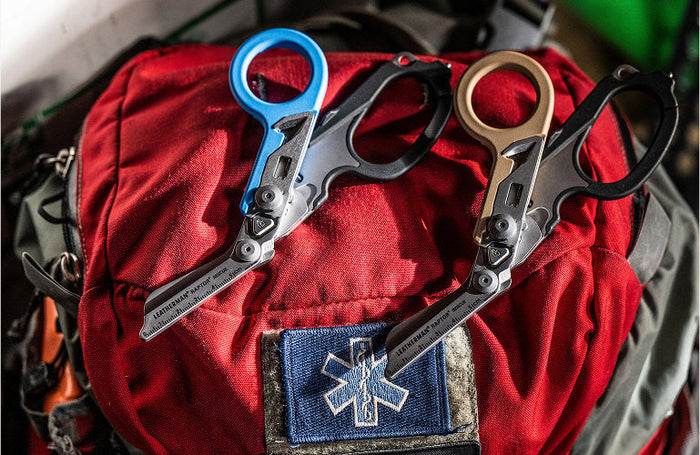 Translation missing: de.Two Leatherman Raptor Rescue shears—one with a blue/blue handle and the other with a tan/black handle—rest on a red first aid backpack adorned with a blue medical star patch, emphasizing their use in emergency medical or rescue situations