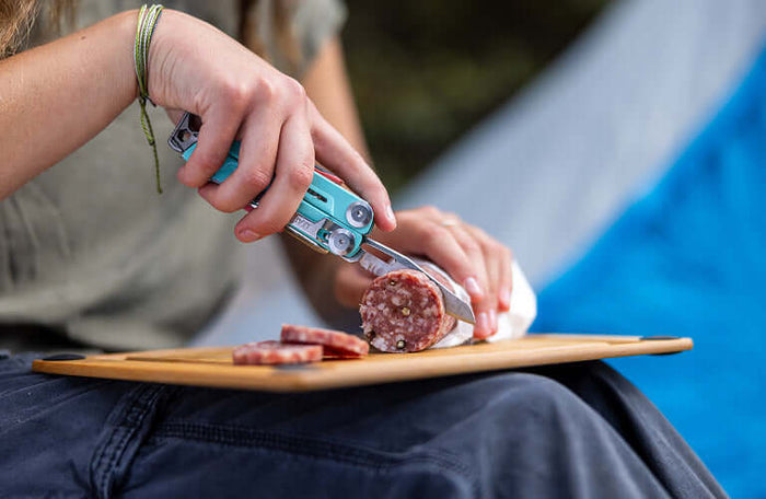 Translation missing: pl.Person using a Leatherman aqua Signal® multitool to slice sausage while camping outdoors