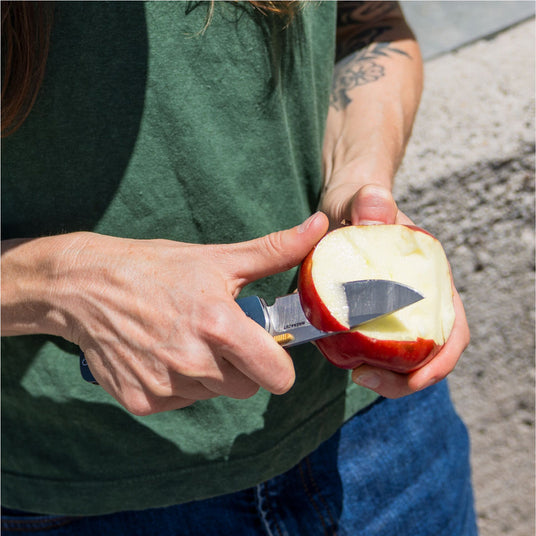 Person using a Leatherman Blazer folding knife to peel an apple outdoors, shown in a close-up with the knife blade slicing through the apple skin.