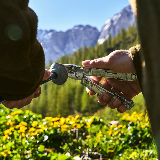 Close-up of a person using a Leatherman Rebar multi-tool to adjust or tighten outdoor gear. The scene is set outdoors with mountains, green pine forest, and yellow wildflowers under a clear blue sky.