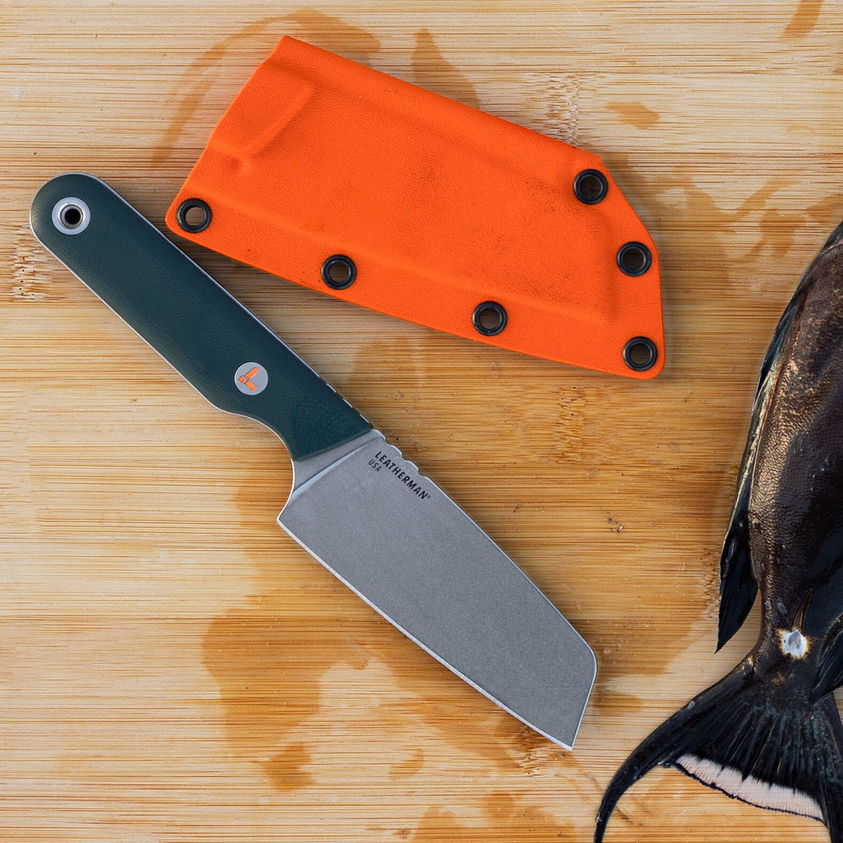 Translation missing: pl.Leatherman Rustle Evergreen knife with a green handle and stainless steel blade resting on a wooden cutting board next to its bright orange sheath and a partially cleaned fish