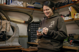 Translation missing: en.A person wearing a brown Leatherman Mr. Wave hoodie standing in a workshop holding a ceramic mug, smiling while surrounded by tools and equipment