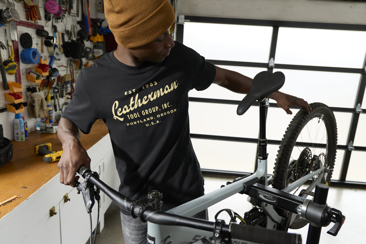 Translation missing: de.A person wearing a black Leatherman Vintage Tee working on a mountain bike in a garage workshop, surrounded by tools and equipment