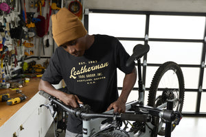 Translation missing: de.A person wearing a black Leatherman Vintage Tee working on a mountain bike in a garage workshop, surrounded by tools and equipment