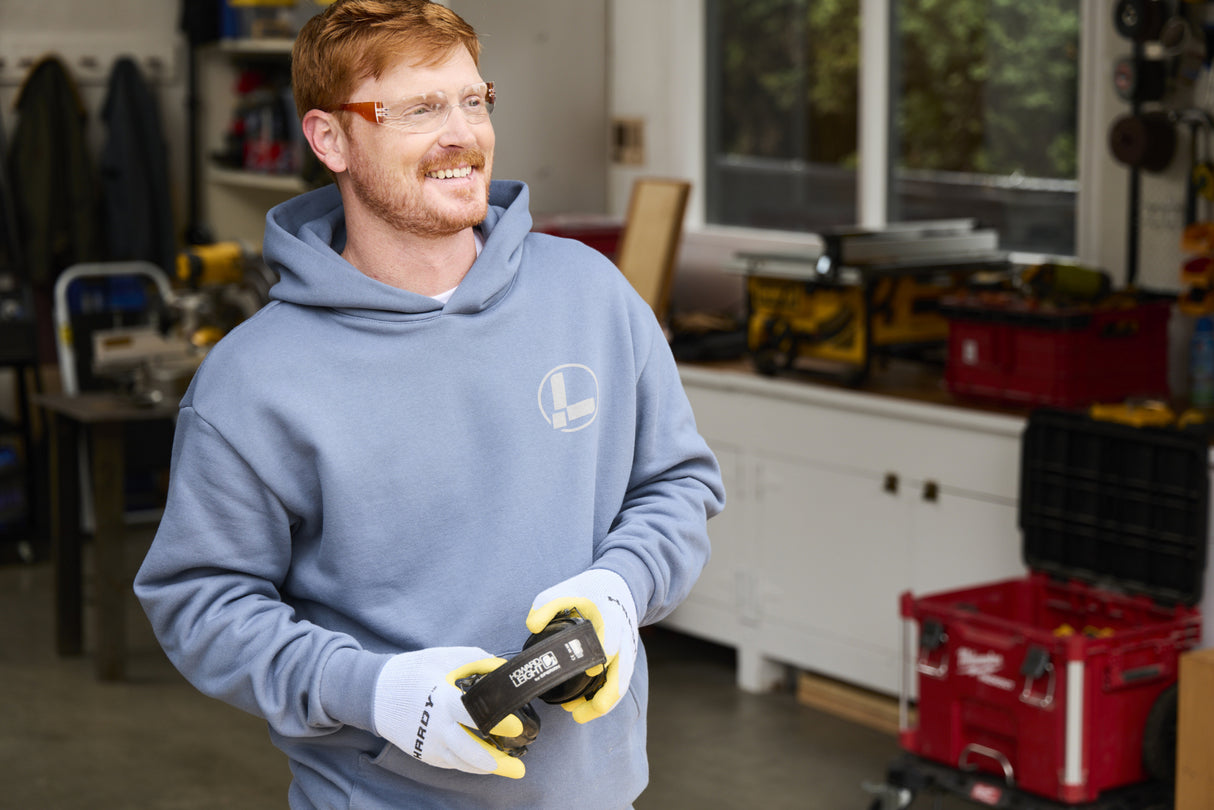 Translation missing: en.A person wearing a storm blue Leatherman Classic Hoodie smiling while holding headphones in a workshop